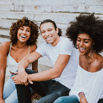 Three smiling friends sit in front of a brick building