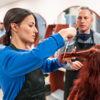 A woman demonstrating a hair cut technique to a man using a comb and her fingers on long red hair