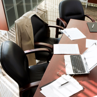 Long wood table covered in documents, pens, and a laptop surrounded by black office chairs